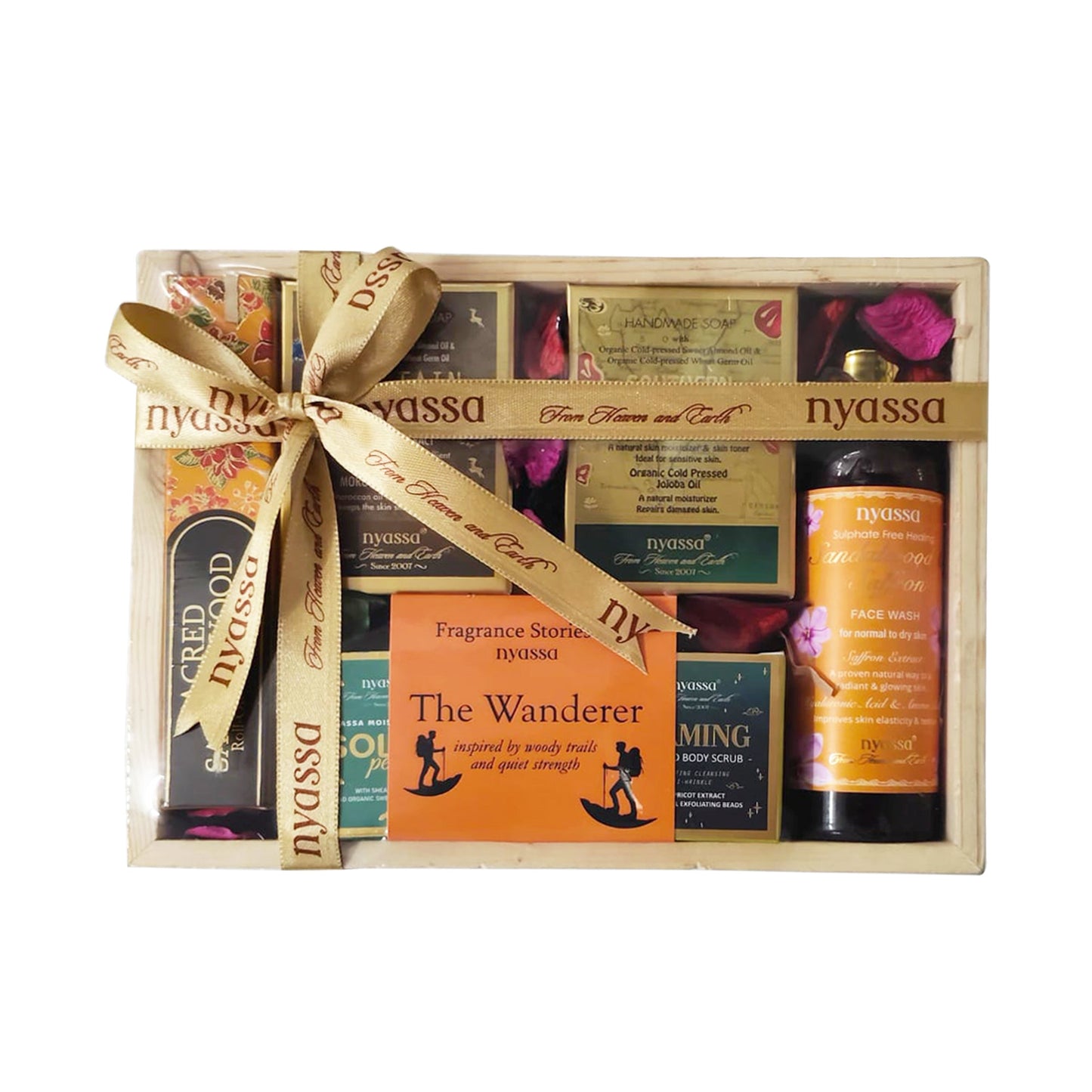 Nyassu gift set in a clear plastic box with a beige wooden base, tied with a beige ribbon printed with the brand name. The set includes a dark amber bottle, an orange bottle, several boxed fragrance products, and decorative rose petals.