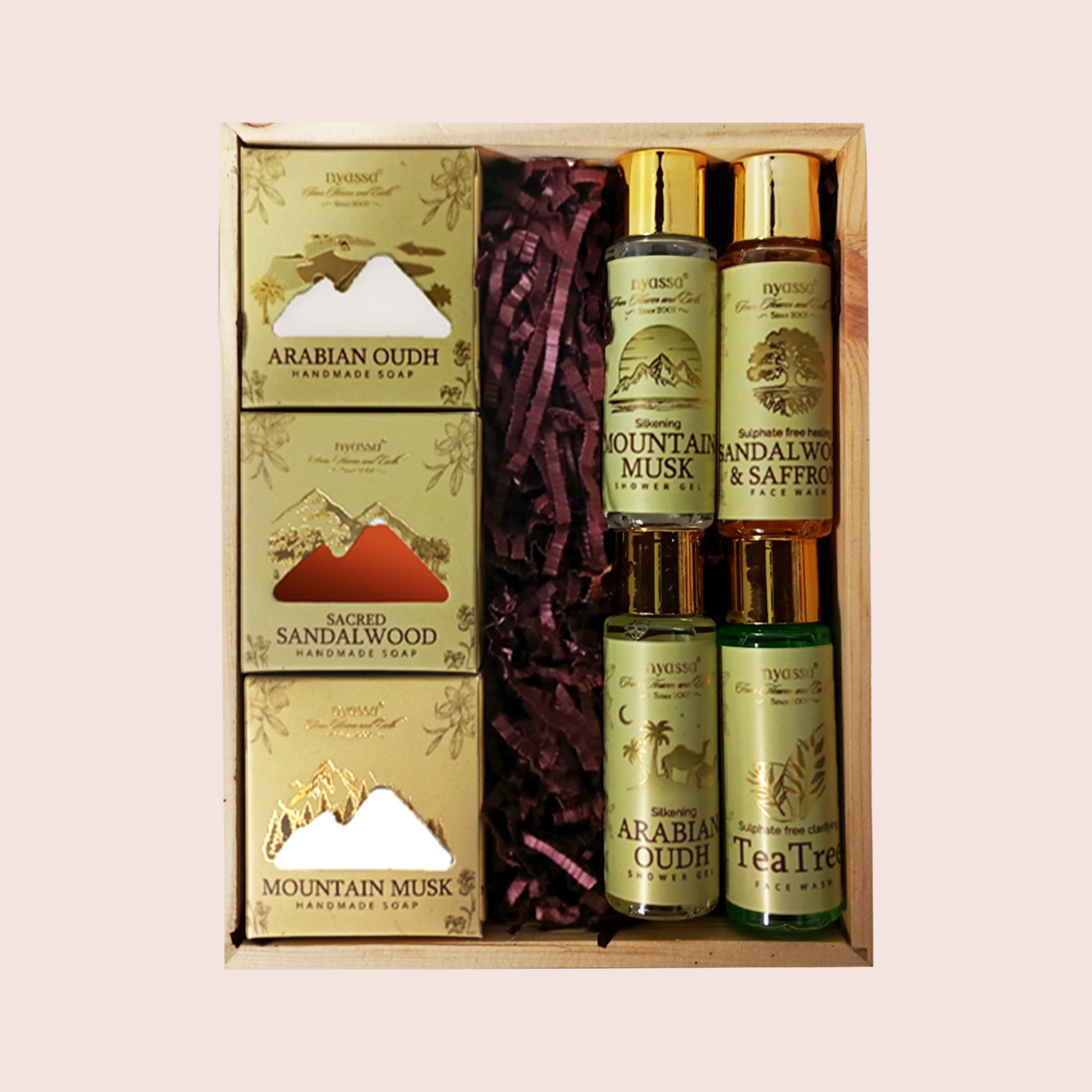 A boxed fragrance gift set containing four boxed Arabian Oudh and Sandalwood scents, two Mountain Musk body mists, and two Tea Tree body mists, arranged in a wooden tray with purple shredded paper filler. The set is branded 'Saffronwood' and photographed against a solid pale pink background.
