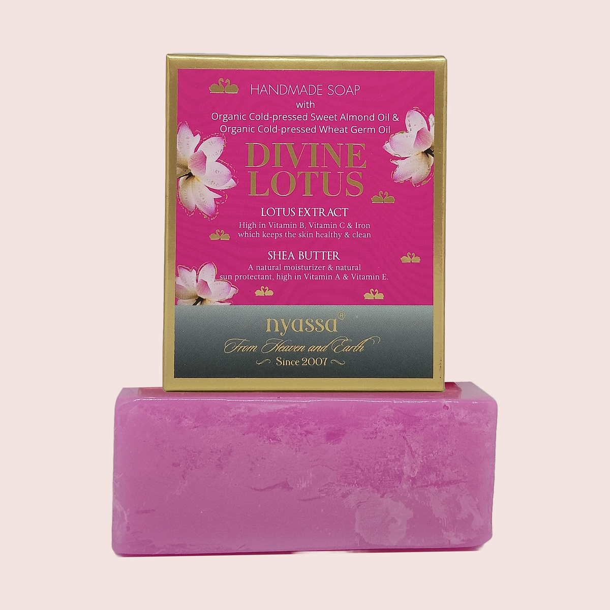 A pink bar of handmade Divine Lotus soap displayed in front of its gold and pink retail box on a light pink background. The box features pink lotus flowers and text highlighting ingredients like lotus extract, shea butter, and almond oil.