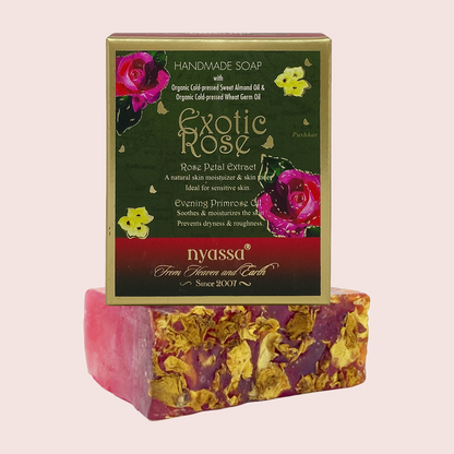 A handmade soap bar with dried flower inclusions sits in front of its branded box. The box is dark green with gold trim and pink/red floral graphics, labeled 'Exotic Rose' by 'nyassa'.