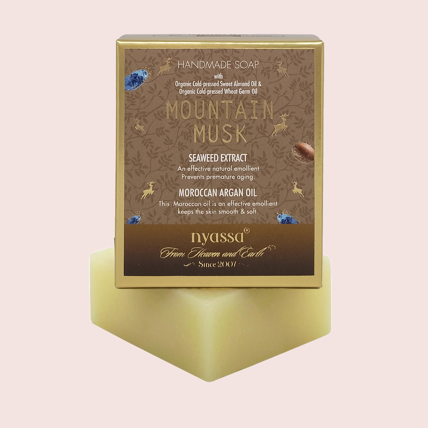 A boxed handmade soap product labeled 'Mountain Musk' by Nyassa, shown standing upright on a pale yellow soap bar against a light pink background. The box is brown with gold text and decorative motifs, and the soap itself is pale yellow.