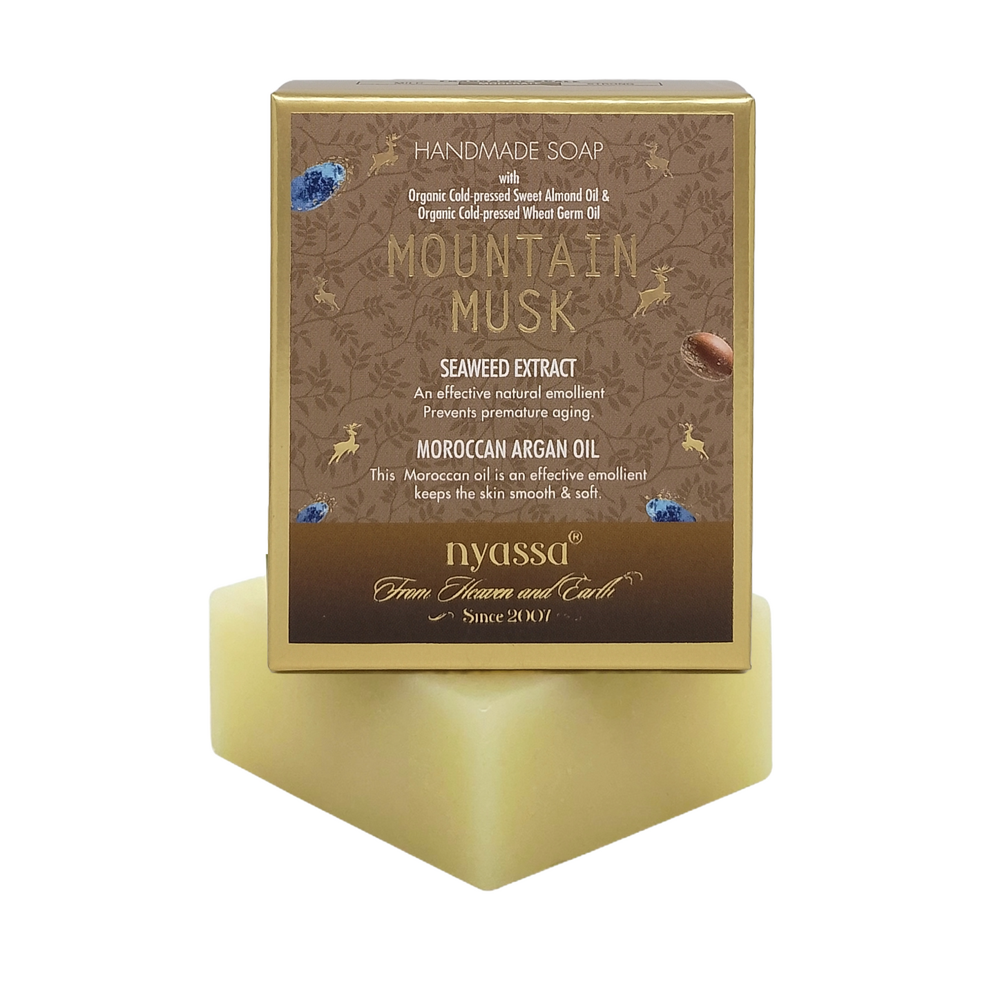 A rectangular bar of handmade soap in a translucent pale yellow cube, displayed on a matching translucent stand. The soap packaging box behind it is brown with gold foil borders and text reading 'Mountain Musk' and the brand 'nyassa'.