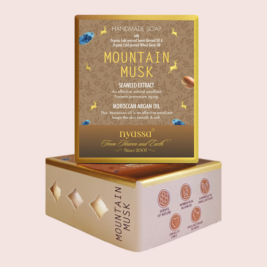 A boxed handmade soap product labeled Mountain Musk, shown with the top box standing upright above a lower box on a pale pink background. The packaging is primarily warm tan and gold with brown text and small illustrated accents, including blue gem-like shapes and a small camel graphic.