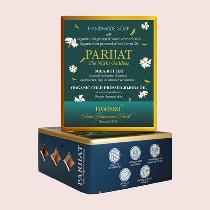 A boxed handmade soap product shown on a plain light pink background. The top box is the front-facing soap package with a dark teal-green label, gold border, white floral illustrations, and the brand name PARIJAT The Night Goddess.