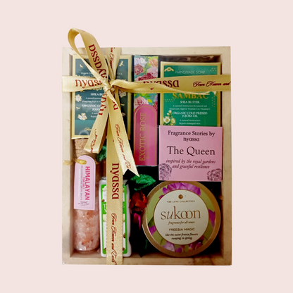 A curated gift box containing fragrance products from Dyssa and Sukoon, tied with a gold ribbon. The box includes a pink bath salt, a green and pink soap bar, a green and pink body butter bar, and a round tin of Freesia Magic diffuser wax.