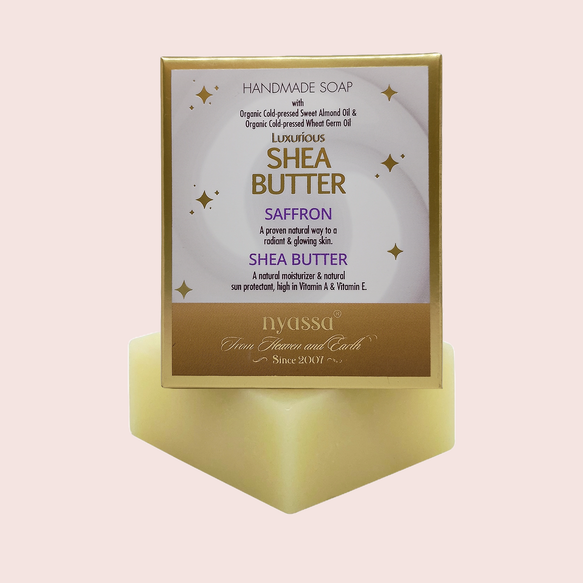 A handmade Shea Butter soap bar in a gold rectangular box with a white and gold label, displayed on a matching gold pedestal against a pale pink background. The label reads 'Handmade Soap with Organic Cold Pressed Sweet Almond Oil & Organic Cold Pressed Wheat Germ Oil LUXURIOUS SHEA BUTTER SAFFRON' and shows the brand 'nyssa'.