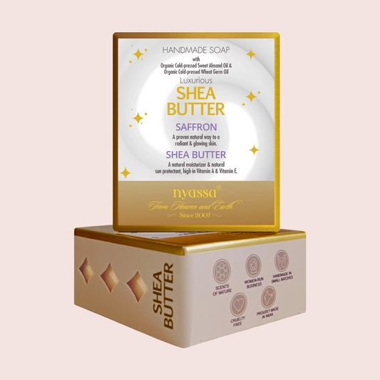 Two stacked rectangular boxes of handmade shea butter soap on a pale pink background. The top box is white with gold and purple text and small gold star graphics, while the bottom box is cream with gold trim and printed icons on the side.