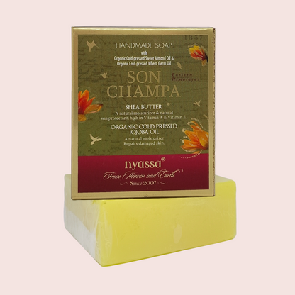 A handmade soap bar wrapped in clear plastic, displayed in front of its product packaging box. The box is olive green with a burgundy bottom band and features the brand name 'Nyassa' and product name 'Son Champa'.