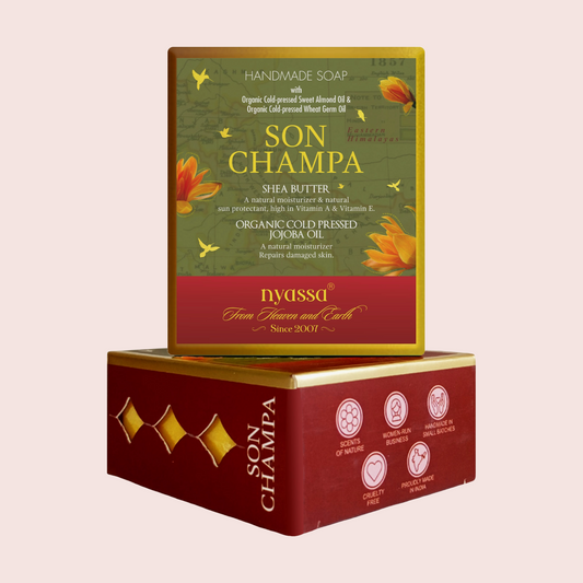 Two stacked rectangular boxes of Nyussa Son Champa handmade soap on a pale pink background. The top box is olive green with gold and red sections, featuring shea butter and jojoba oil text and orange flower illustrations.