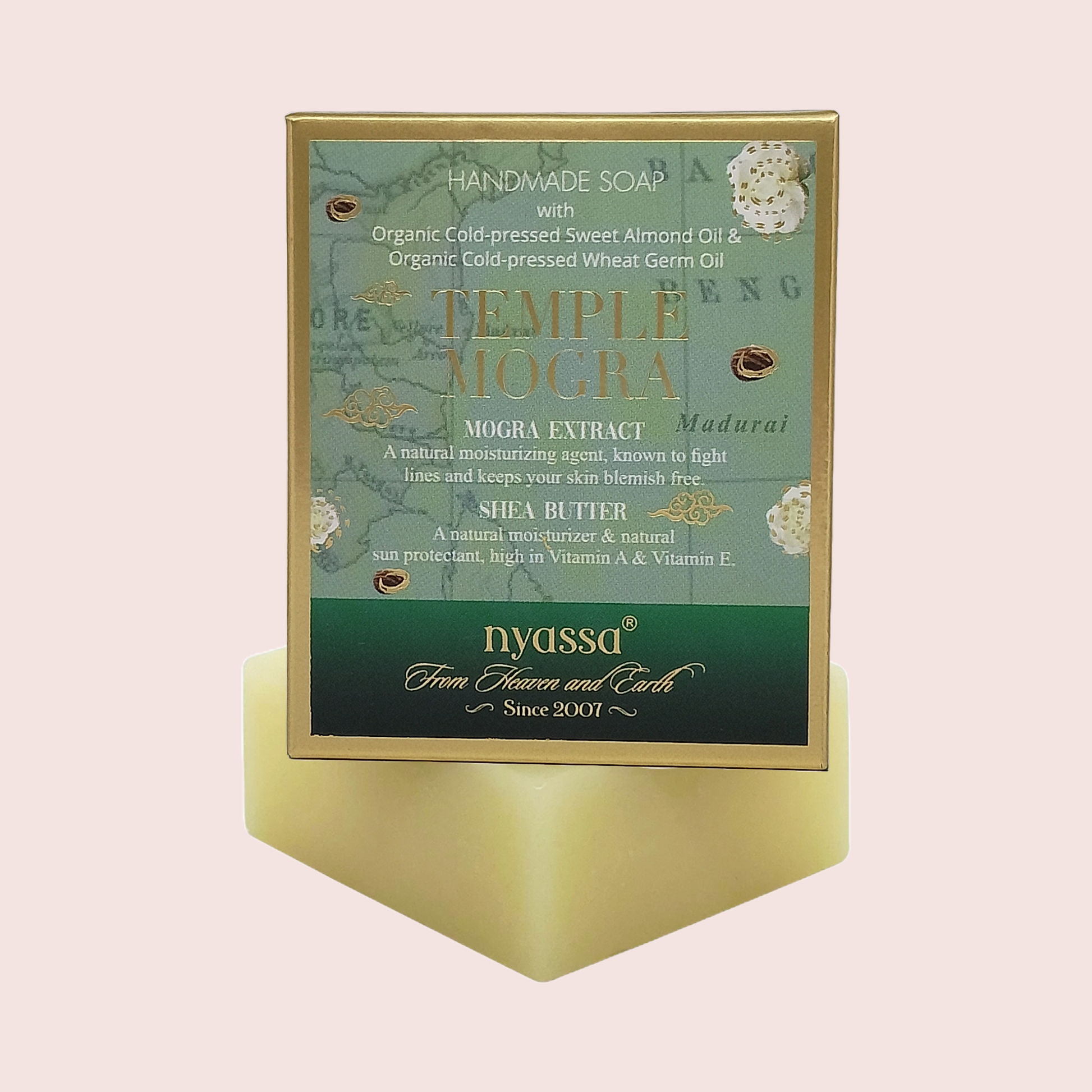 A boxed handmade soap product labeled 'Temple Mogra' by Nyassa, shown standing upright on a pale yellow pedestal against a soft pink background. The front panel is teal and green with gold text and decorative botanical illustrations.