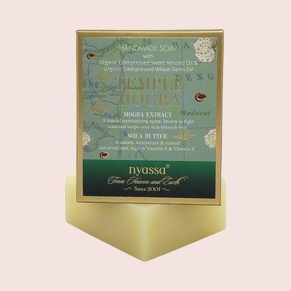 A boxed handmade soap product labeled 'Temple Mogra' by Nyassa, shown standing upright on a pale yellow pedestal against a soft pink background. The front panel is teal and green with gold text and decorative botanical illustrations.