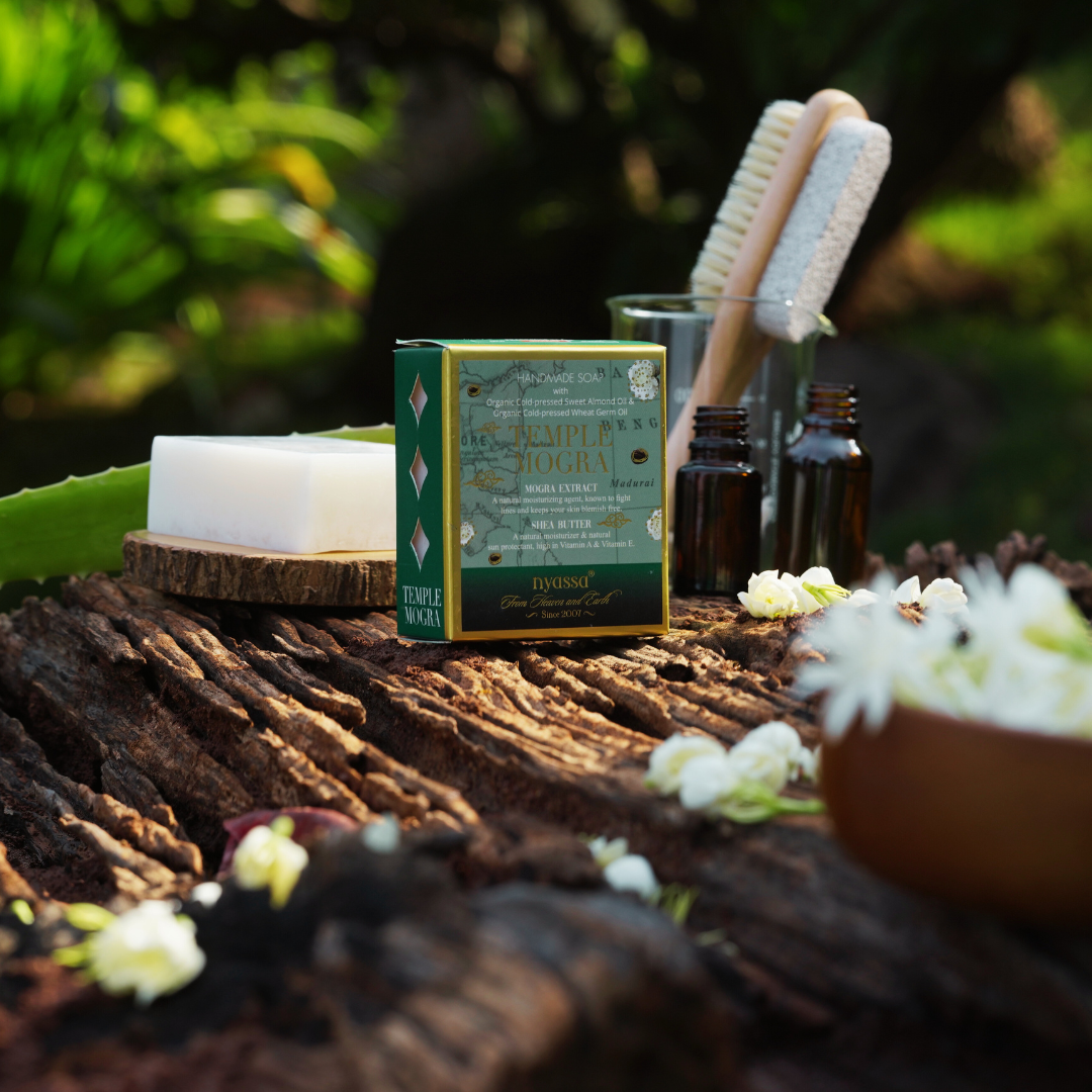 A boxed bath and body set labeled 'Lavender & Jasmine' sits on a rustic log outdoors, styled with a bar of soap, two small amber bottles, two wooden brushes, and white flowers against a blurred green garden background.