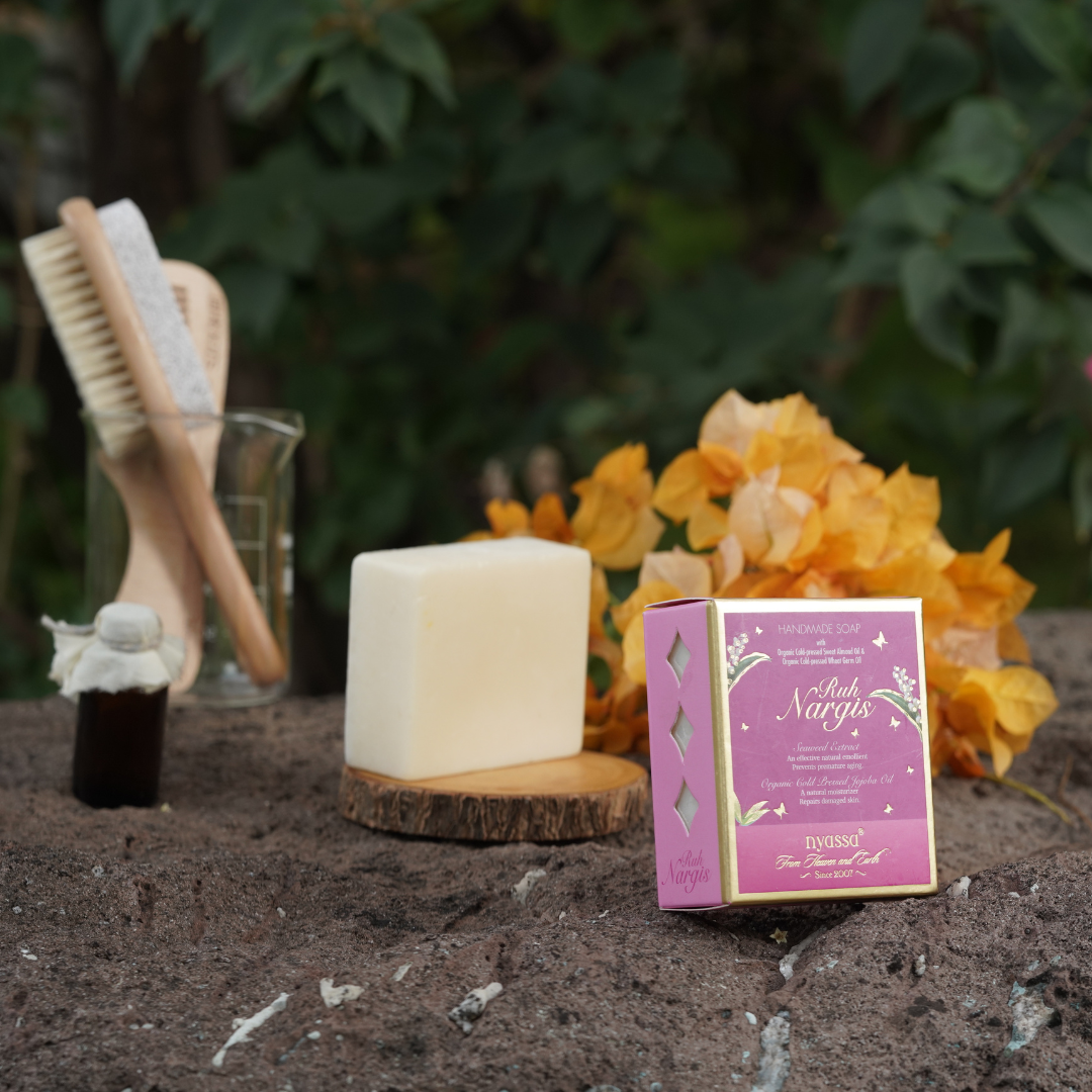 A bar of soap and its decorative box displayed outdoors on a stone surface, surrounded by yellow flowers, wooden brushes, and a small bottle against a blurred green foliage background.