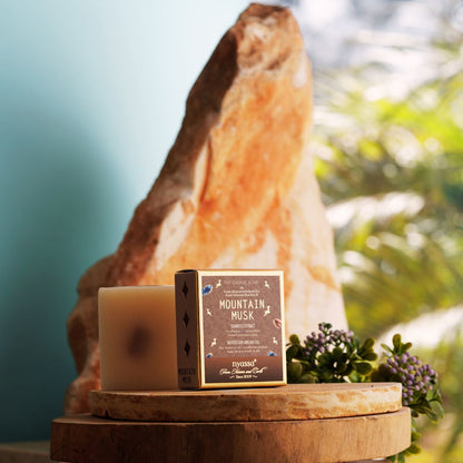 A candle and its box displayed on a wooden pedestal, with a large rock and blurred greenery in the background. The box reads Mountain Musk and shows the brand Myrdas.