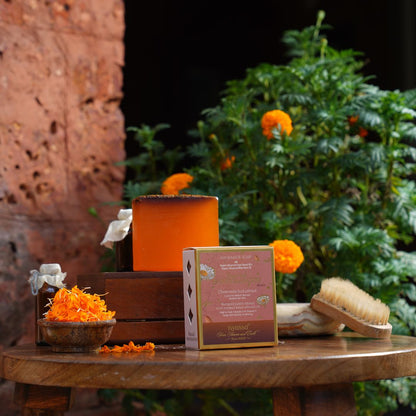 A boxed natural hair product displayed on a wooden table with orange flower petals, a wooden brush, and a green leafy plant in the background. The box is labeled 'pyra.
