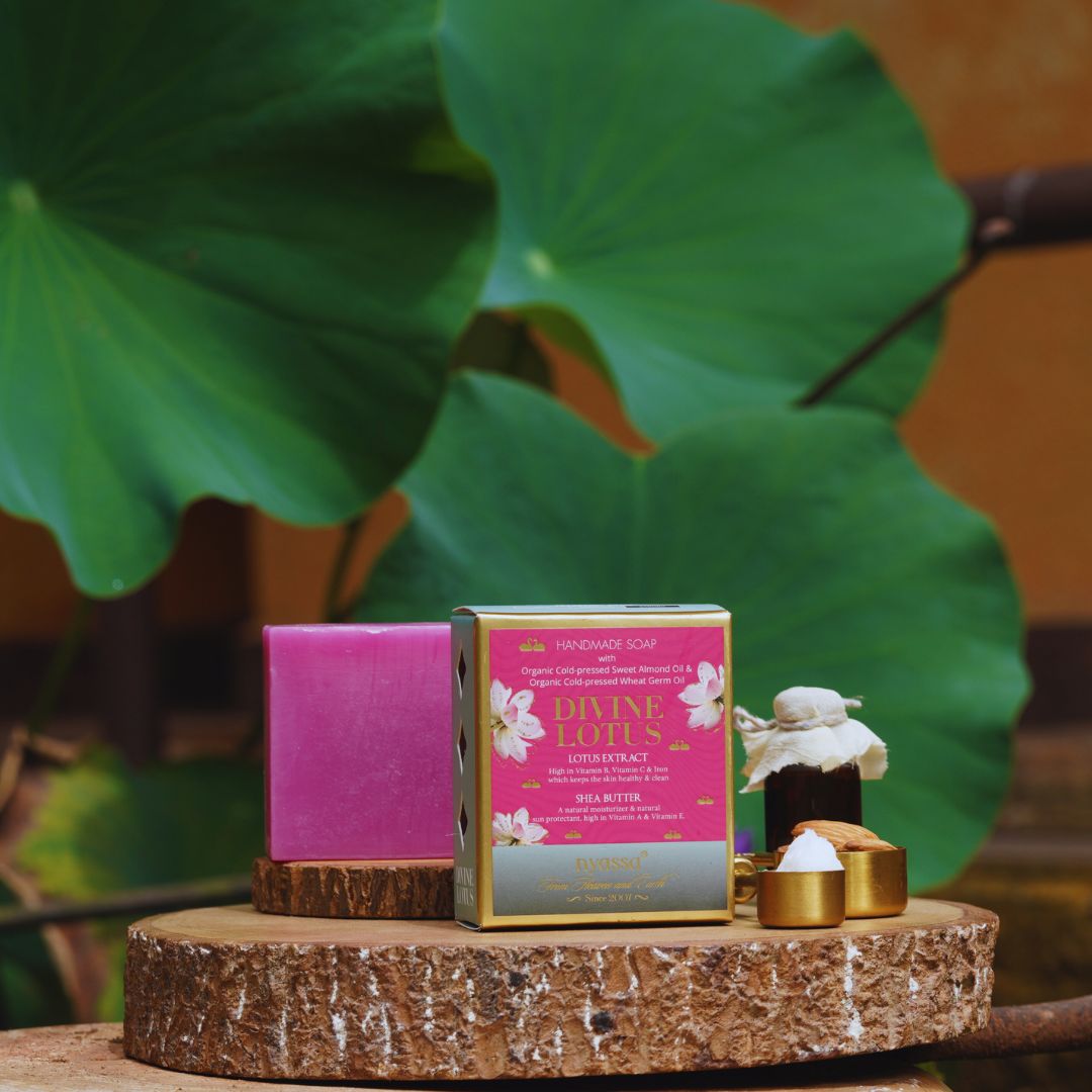 A boxed handmade soap product displayed on a rustic wood slice pedestal, with a pink soap bar beside it and a small bottle with white wax and gold caps in front. Large green lotus leaves and a warm brown background are visible behind the product.