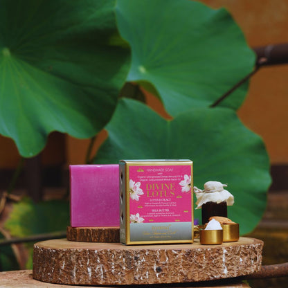 A boxed handmade soap product displayed on a rustic wood slice pedestal, with a pink soap bar beside it and a small bottle with white wax and gold caps in front. Large green lotus leaves and a warm brown background are visible behind the product.