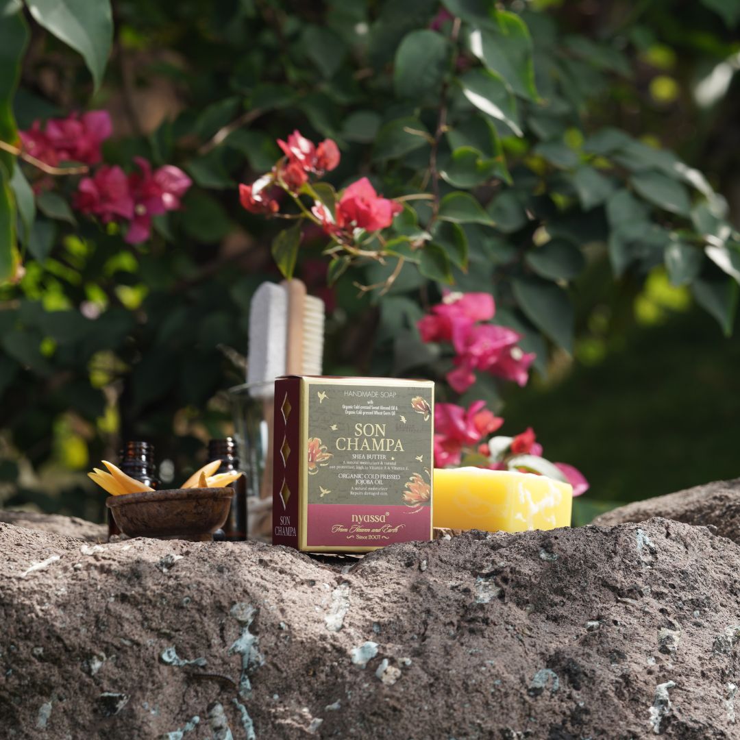 A boxed product labeled 'SON CHAMPA' sits on a rough stone outdoors, surrounded by green foliage, pink flowers, a yellow bar of soap, small dark bottles, and a wooden spoon with yellow sticks. The scene is styled in a natural garden-like setting, so this is Lifestyle rather than Product/Packaging.