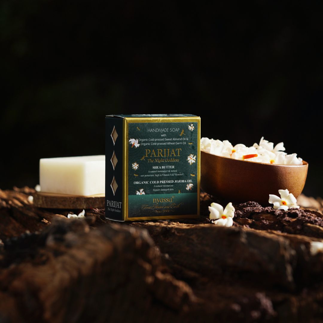 A boxed bar of Parijat Marigold handmade soap stands on a rustic wooden surface, with a lit candle and a bowl of white marigold flowers nearby against a dark, moody background.