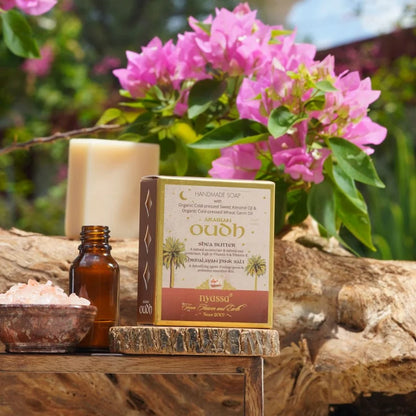 A boxed handcrafted soap product labeled 'Oooh' and 'Nyussa' stands on a rustic wood slice outdoors, with pink bougainvillea flowers, a candle, a small amber bottle, and a bowl of bath salts in the background.