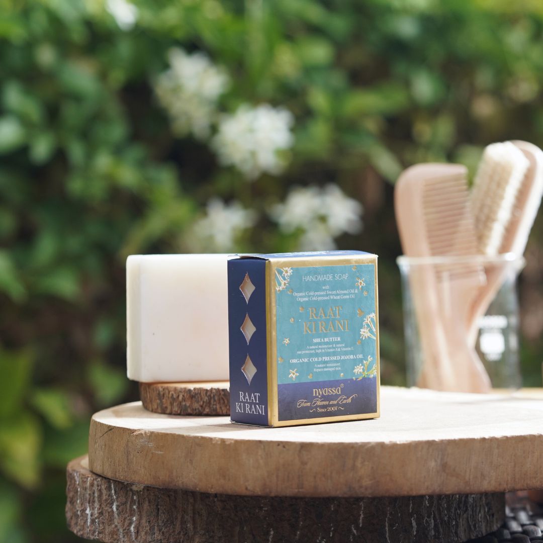 A boxed bath and body set sits on a rustic wooden table outdoors, with a white bar of soap behind it and wooden comb-like tools in a glass container blurred in the background. The product box is blue and navy with gold decorative text reading 'RAAT KURANI' and 'DIVINS'.