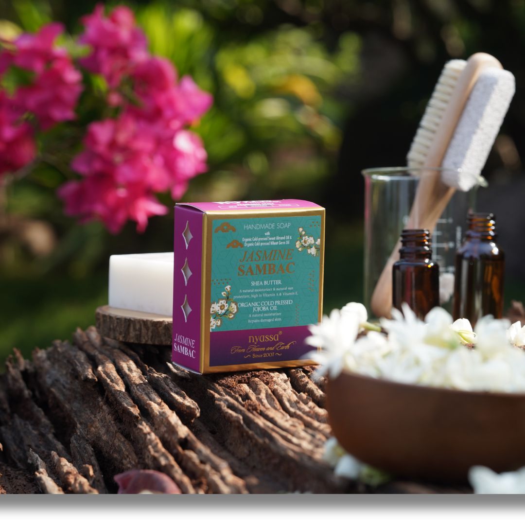 A boxed bath product labeled 'Jasmine Sambac' and 'INVOSSO' sits on a rustic log outdoors, with a white candle, amber bottles, a glass brush holder, and white flowers in the background.