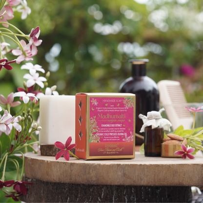 A boxed bath product labeled 'Mochhwaladi' sits on a rustic wooden surface outdoors, surrounded by flowers, a candle, a dark bottle, and a comb, with a blurred green garden background.