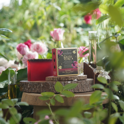 A boxed candle product labeled 'Exotic Rose' sits on rustic wood slices outdoors among green leaves and pink roses. A red translucent candle block is placed beside the box, with small glass vessels in the background.