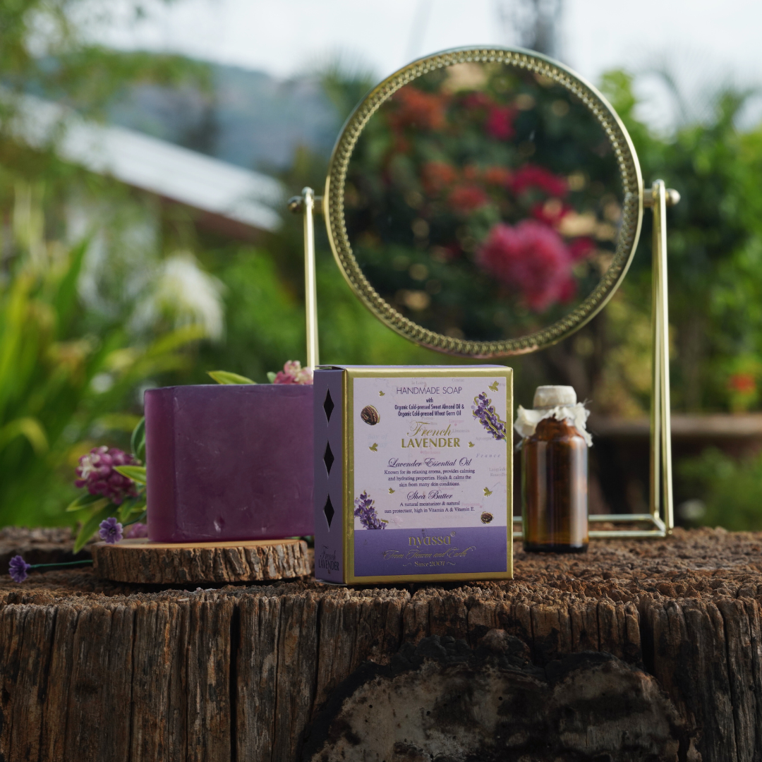 Lavender incense soap and oil set displayed on a rustic tree stump with a gold-framed mirror, featuring a purple wax block, lavender-themed box, and amber oil bottle against a blurred garden background.
