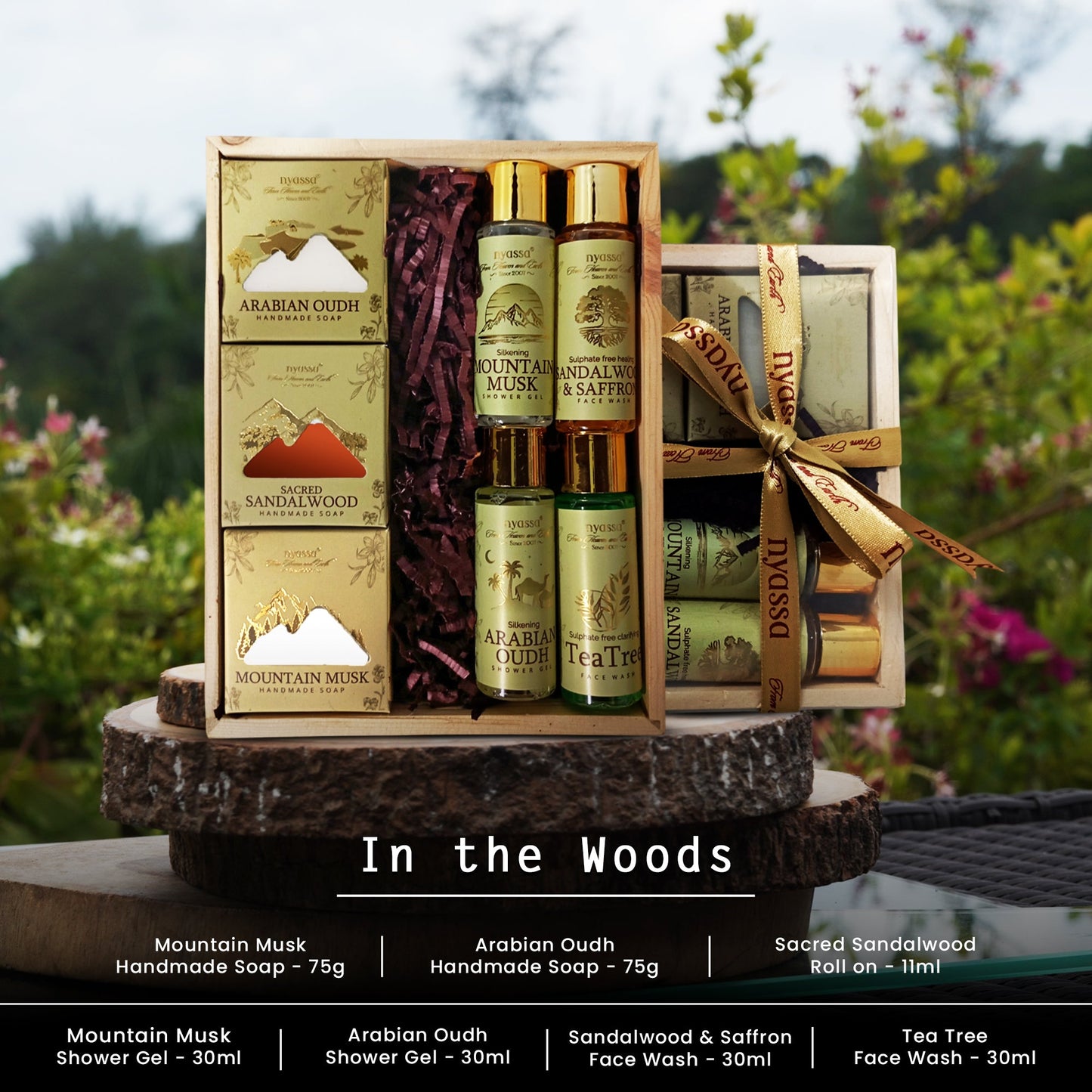 A boxed bath and body gift set displayed outdoors on a wooden stump, with the set opened to show packaged items and three small bottles. The set includes Mountain Musk Handmade Soap, Arabian Oudh Handmade Soap, Sacred Sandalwood & Saffron Face Wash, and Tea Tree Face Wash.