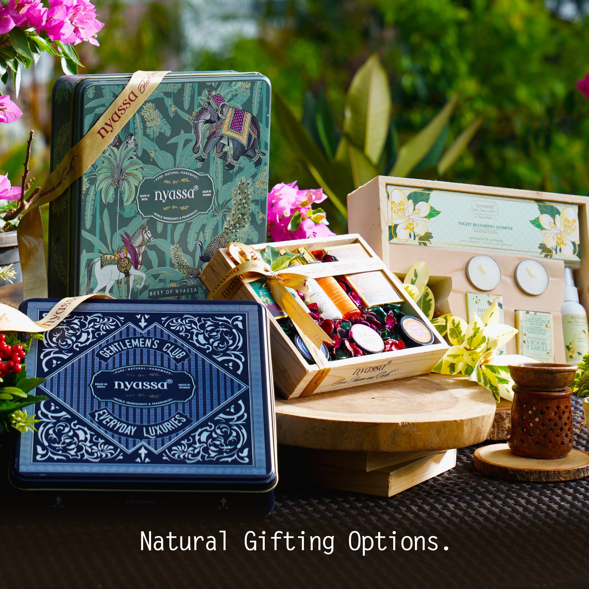A styled gift set display featuring multiple Pycussa branded items, including a teal illustrated tin, a navy blue decorative box, an open wooden box with assorted chocolates and bars, and other gift containers, arranged on wooden stands with flowers and greenery in the background.