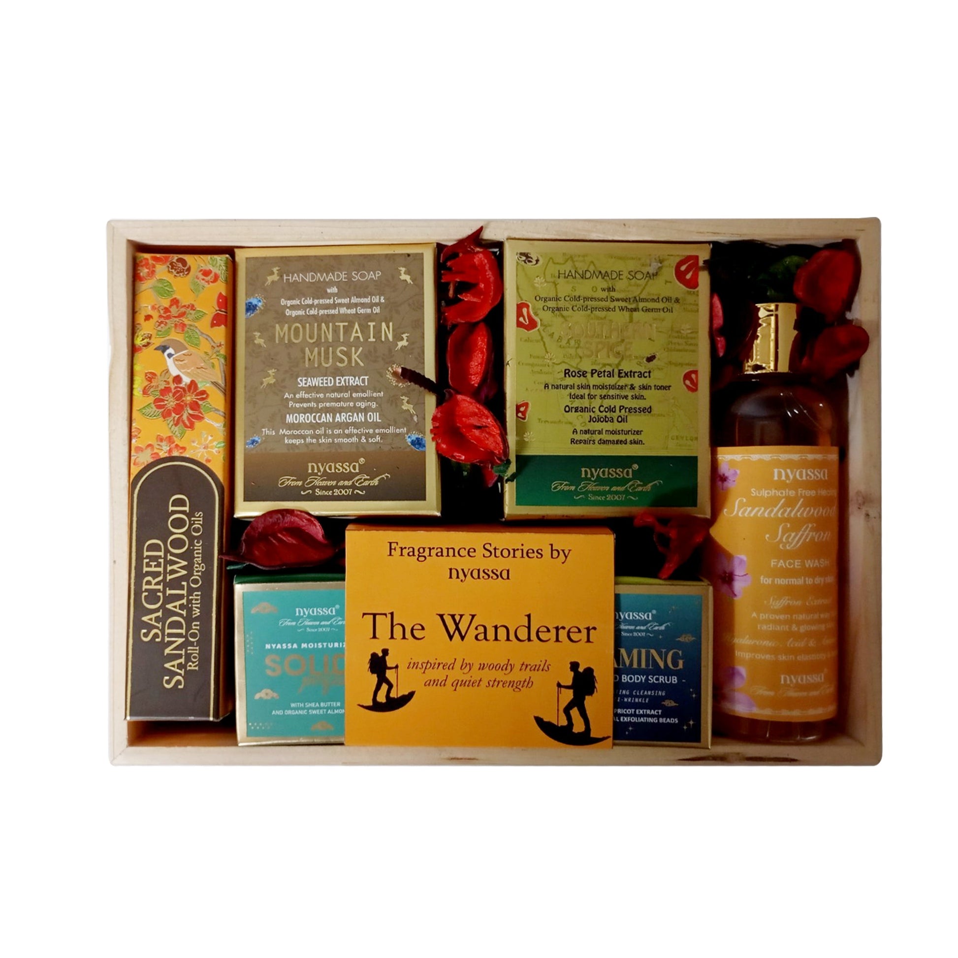 A curated gift set in a light wood tray, featuring products from the brand nyssa, including Sacred Sandalwood body oil, Mountain Musk handwashed soap, The Wanderer fragrance, and a body scrub, decorated with red rose petals.